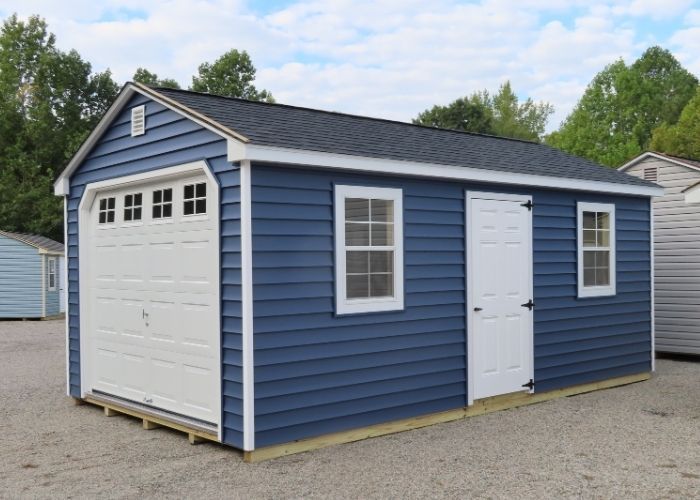 Dark blyue detached garage with white trim, a garage door, side door, and window on gravel driveway