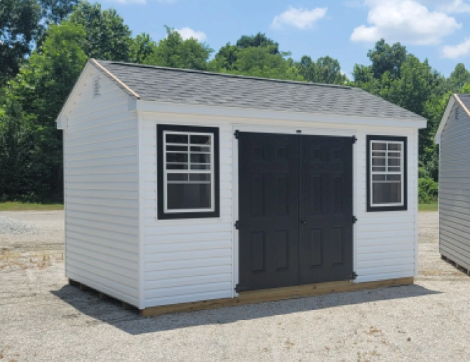 Small white storage shed with black double doors and windows on a gravel lot.