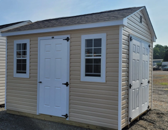 Small beige shed with white trim, windows, and double doors on a gravel lot.
