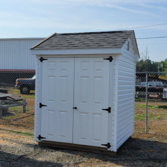 Small white outdoor storage shed with double doors and black hinges in a fenced yard under a blue sky