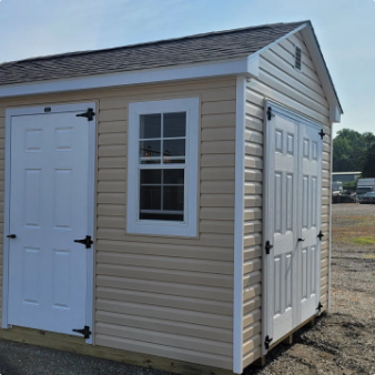 Beige storage shed with white doors and window, sitting outdoors on gravel.