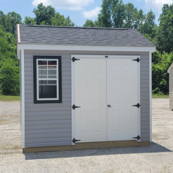 Small gray shed with white double doors and a black-trimmed window on a gravel lot.
