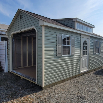 Small light-green shed with open garage-style front, white door and windows, on a gravel lot.