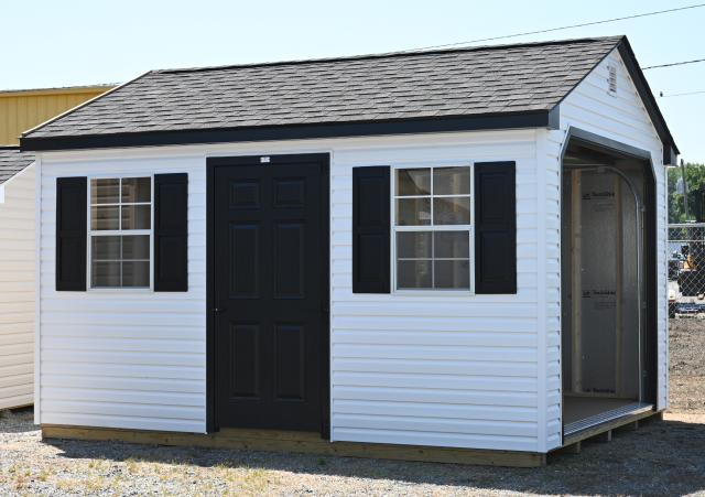 Small white shed with black door and shutters, shown outdoors on gravel.