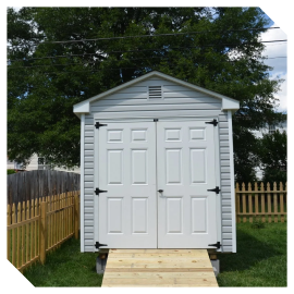 White backyard storage shed with double doors and a ramp, surrounded by a wooden fence and trees.
