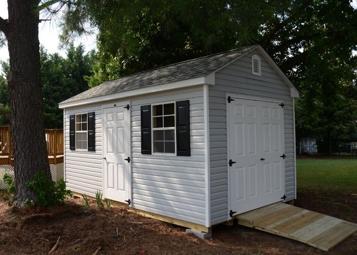 Heritage gray storage shed with black shutters and double doors beside a large tree in a wooded yard