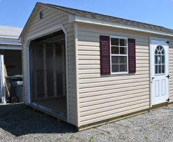 Small beige shed with maroon shutters and a white door, shown on a gravel lot.