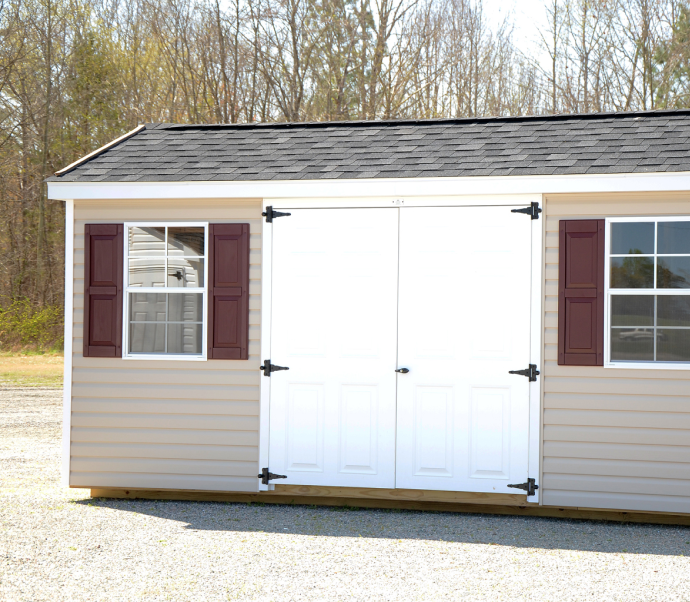 Small beige shed with white double doors and maroon shutters on a gravel lot