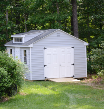 Gray storage shed with white double doors in a grassy yard surrounded by trees