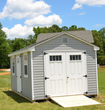 Small gray garden shed with white double doors and a ramp on a grassy lawn under a blue sky.