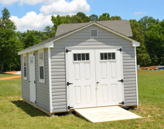 Small gray shed with white double doors and a ramp on a grassy lawn under a blue sky