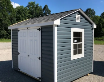 Gray storage shed with white trim, double doors, and a window on a gravel lot