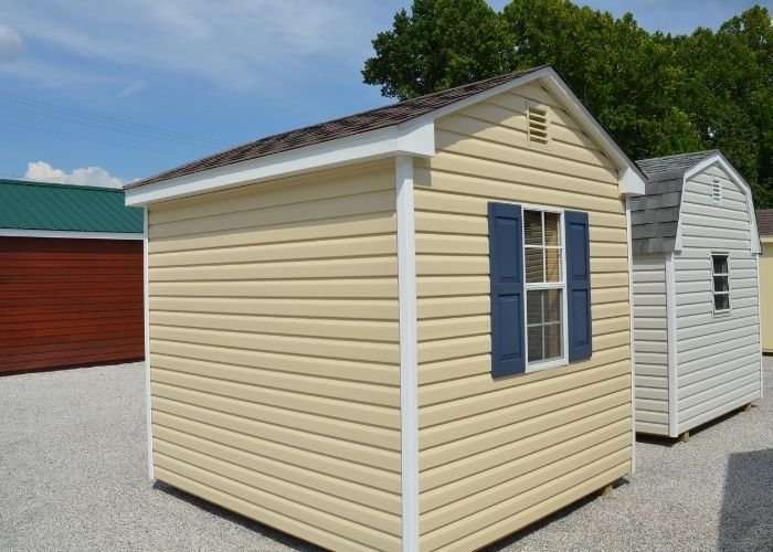 Prairie wheat shed with white trim and blue shutters on a gravel lot, with other sheds nearby