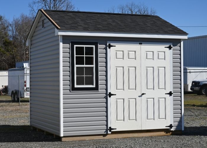 Slate gray shed with white double doors and a window, standing outdoors on a gravel lot.