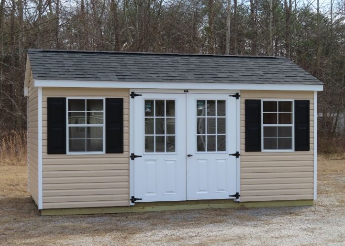 Tan shed with black shutters and double white doors on a gravel lot, with trees in the background