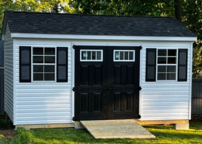 White garden shed with black double doors and shutters on a lawn, viewed from the front.