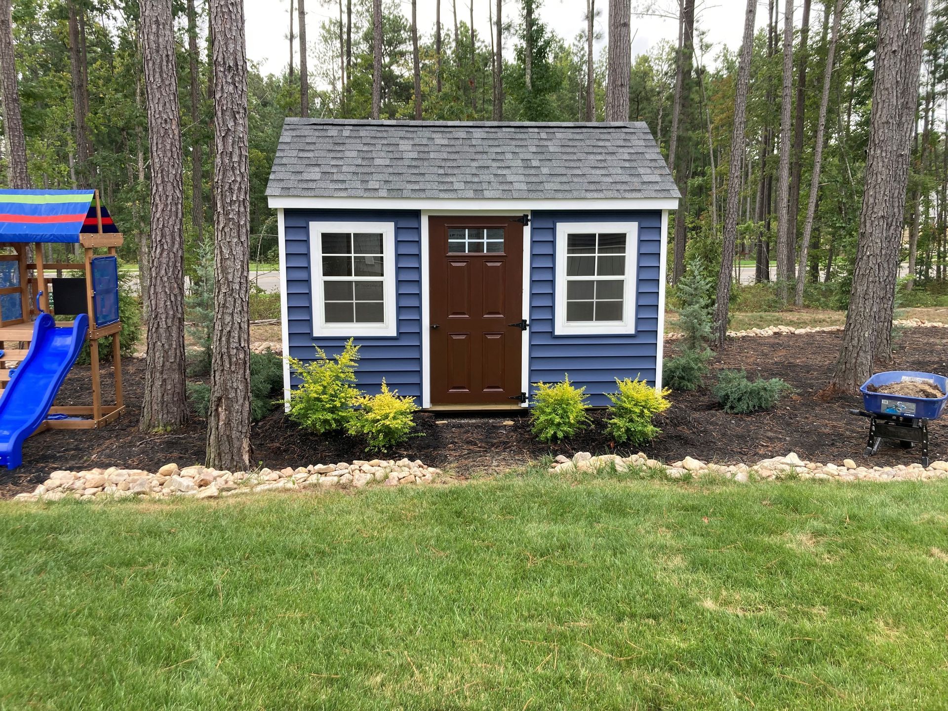 Blue backyard playhouse with white trim and brown door in a wooded yard, beside grass and a slide