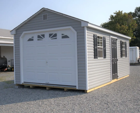 Gray garage-style storage shed with white trim and double doors on a gravel lot