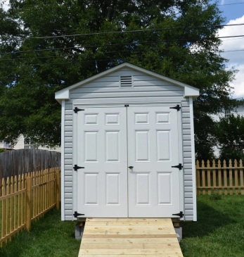Gray backyard storage shed with double doors and a wooden ramp, framed by a fence and trees.