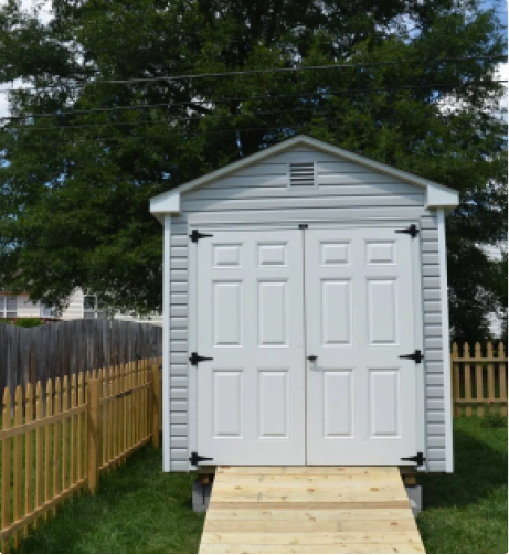 Small white backyard storage shed with double doors and a wooden ramp, surrounded by a fenced lawn.
