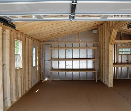 Empty unfinished room with exposed wood framing, open garage door, and built-in shelves against the back wall