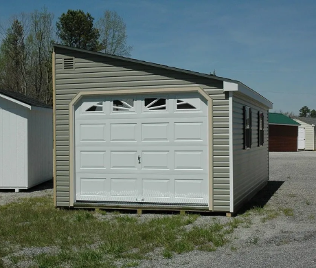 Small beige detached garage with white double doors on a gravel lot under a clear sky