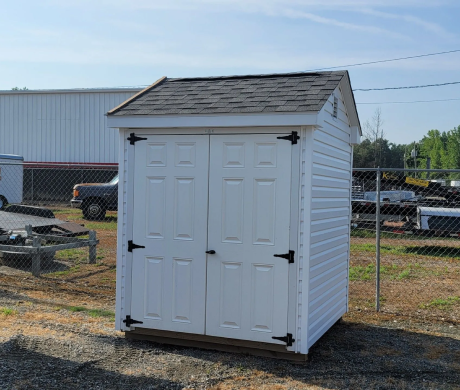 Small white storage shed with double doors and a gray shingled roof on a gravel lot outdoors