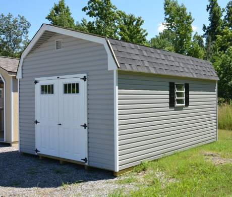 White vinyl shed with double doors and a small window, sitting on gravel beside a house and trees.