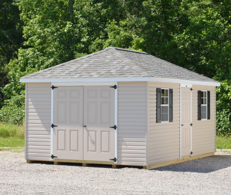 White vinyl storage shed with double doors and windows on a gravel lot, surrounded by trees