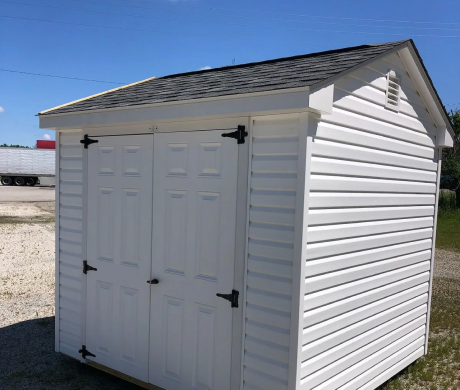 White storage shed with double doors and gray roof on a gravel lot