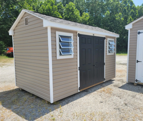 Small beige storage shed with white trim, dark double doors, and two windows on a gravel lot
