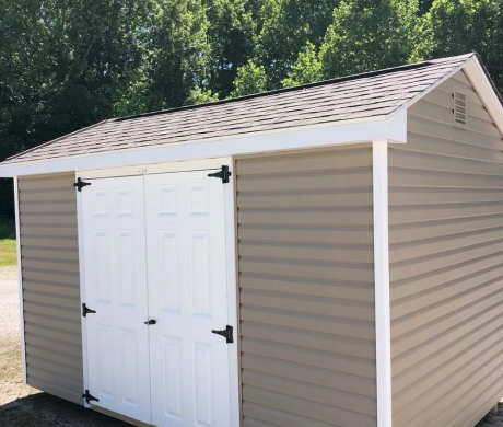 Small beige storage shed with white double doors and black hinges, set outdoors by trees