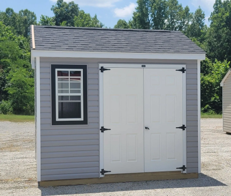 Small gray shed with white double doors and a window, sitting on a gravel lot near trees.