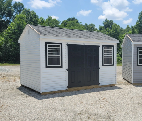 Small white storage shed with black double doors and windows on a gravel lot under a blue sky
