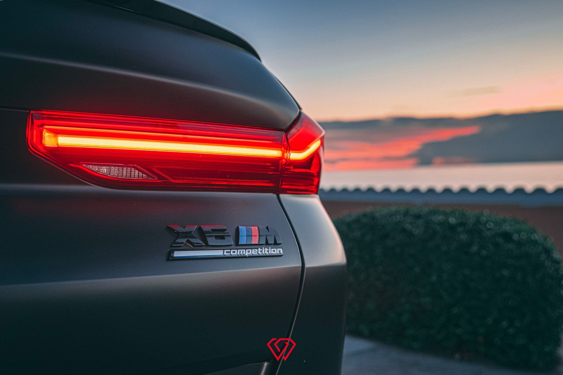 Rear view of a black BMW X5 M SUV with illuminated taillight at dusk, near water and a bush.
