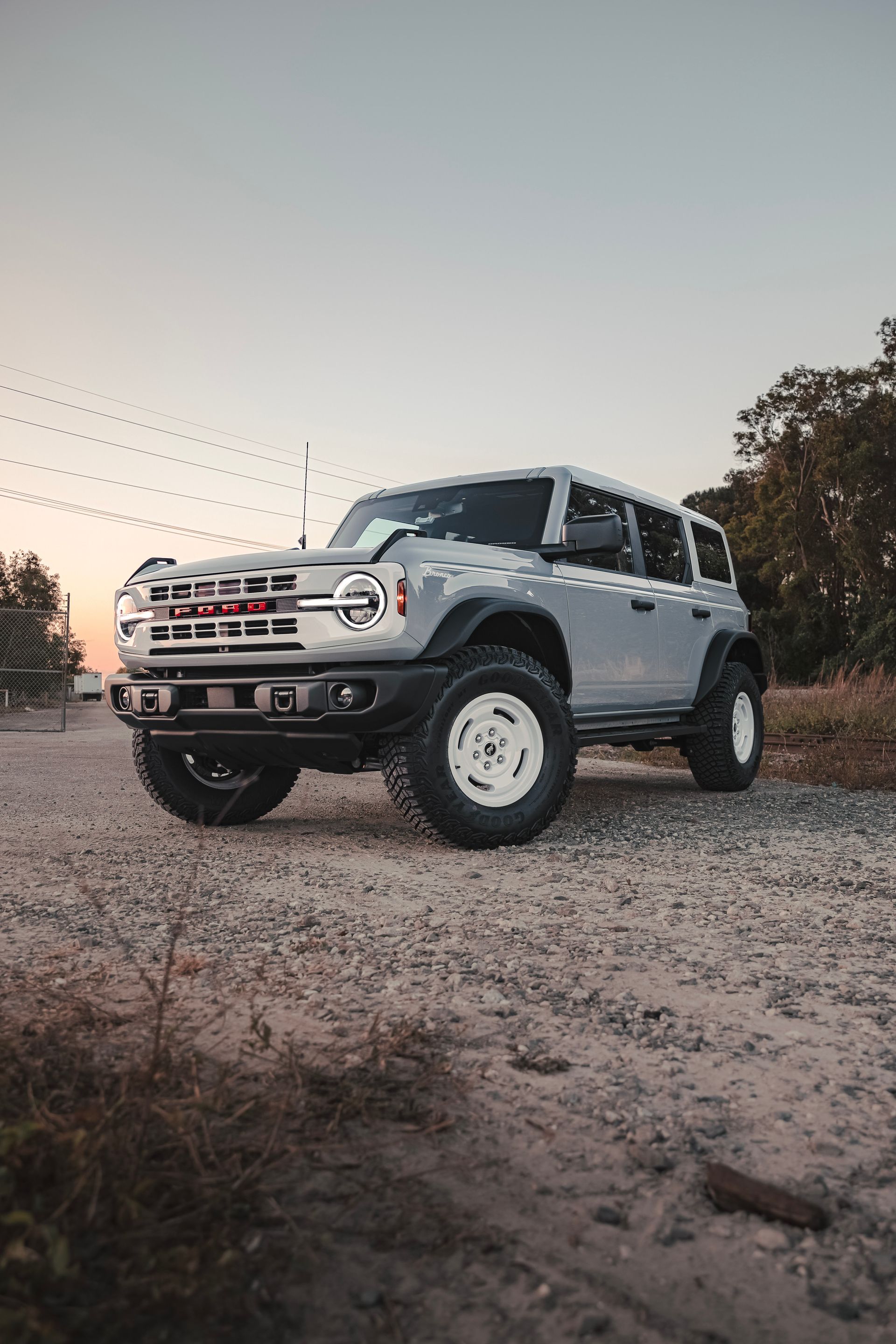 A light gray Ford Bronco with white wheels parked on a dirt road at sunset.