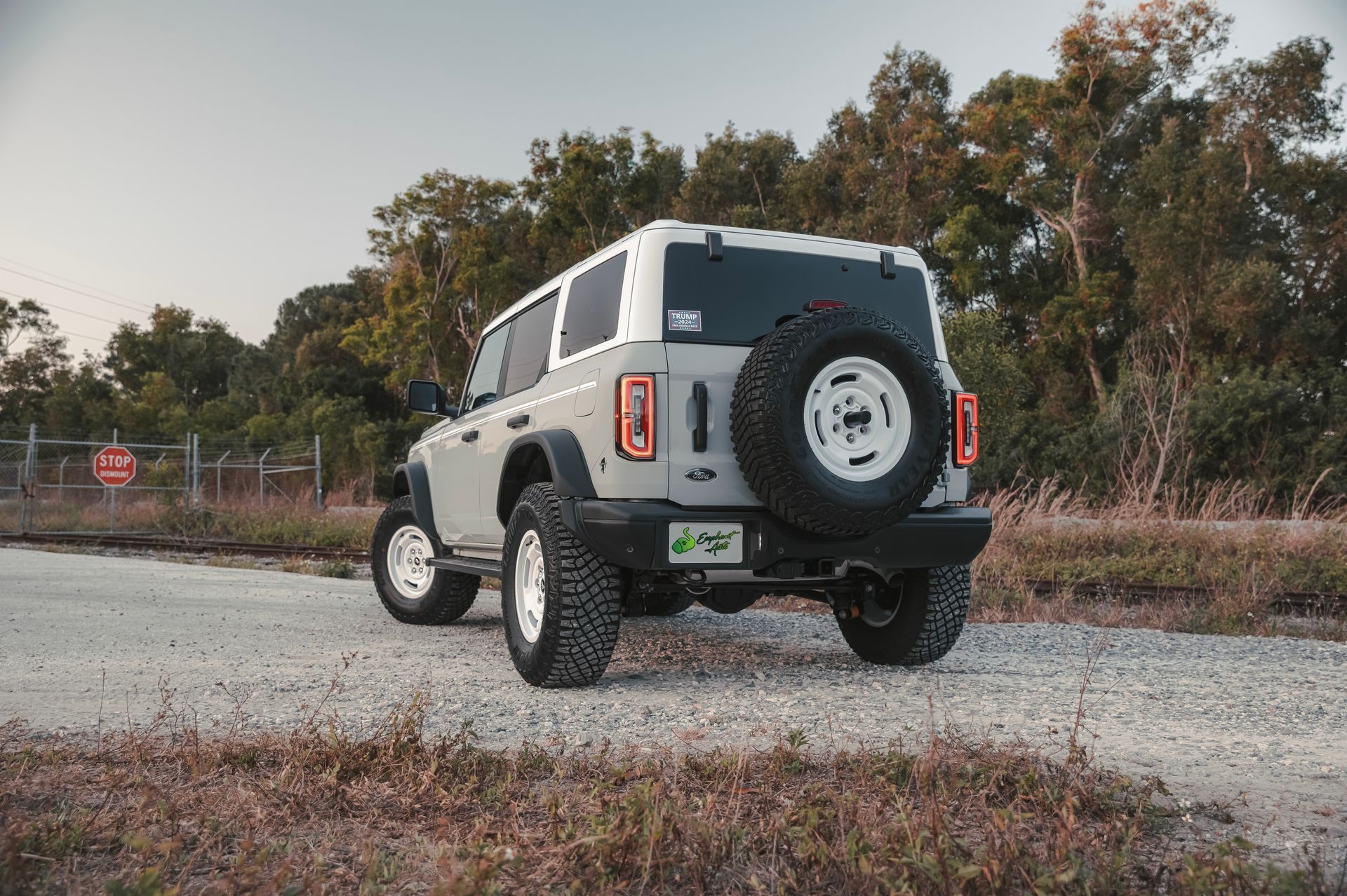 Grey Ford Bronco with white top, parked on gravel road with spare tire.