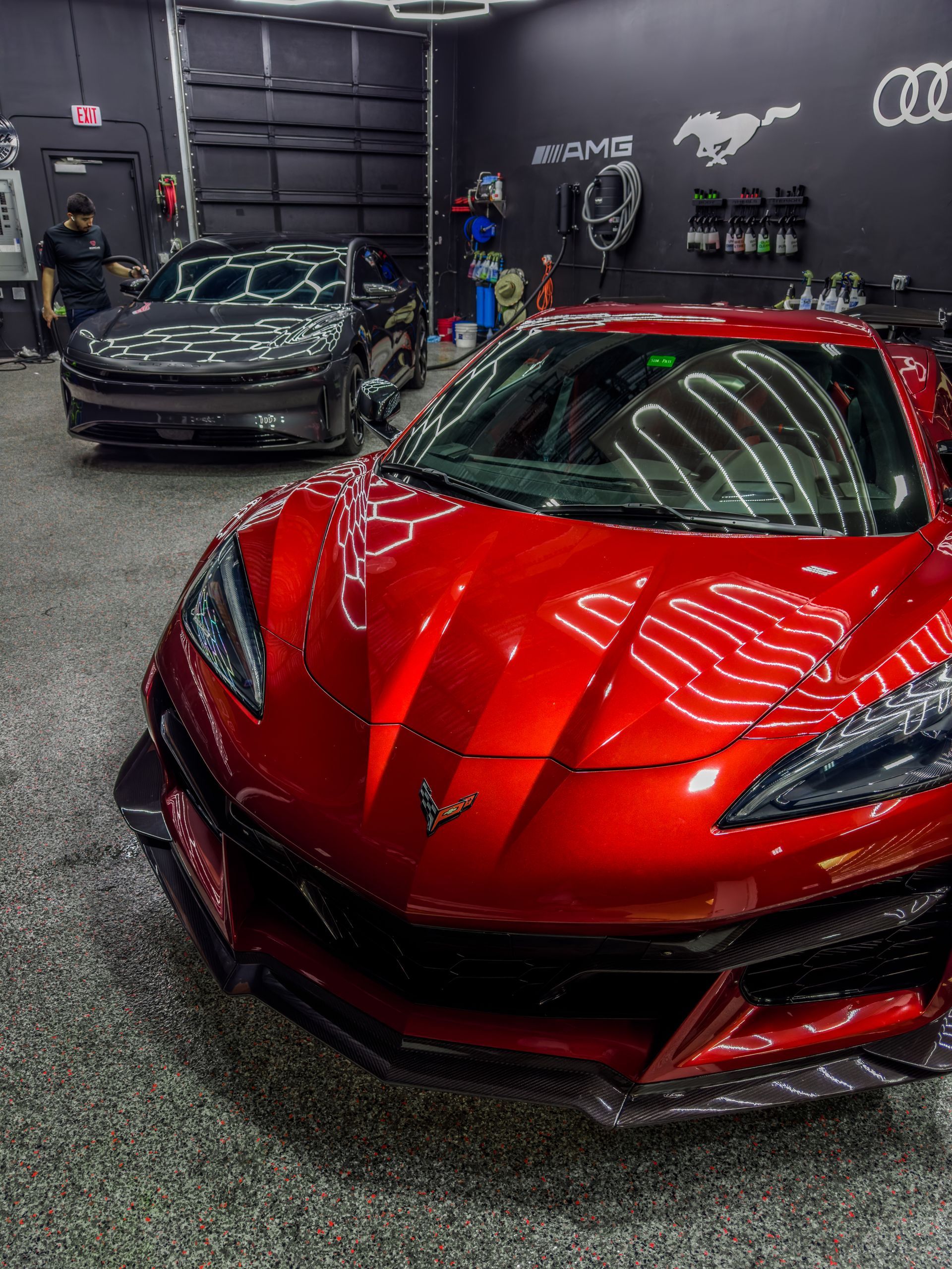 Red sports car in a garage, with a black car in the background. The garage has professional detailing tools and supplies.