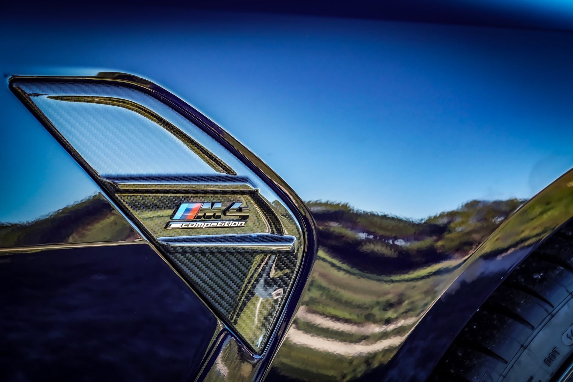 Close-up of a blue BMW M4 side vent with carbon fiber accents, reflecting sky and landscape.