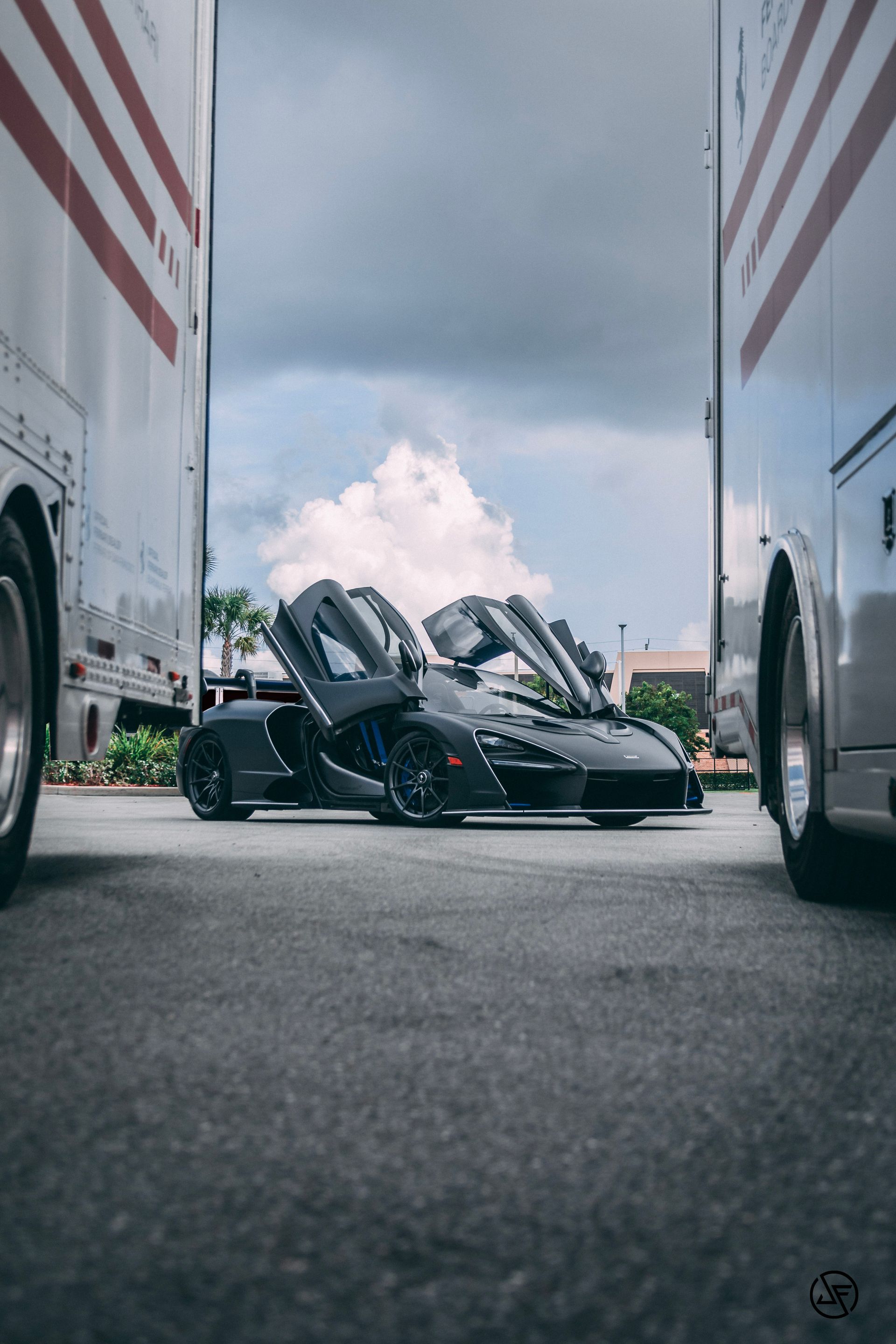 Black McLaren sports car with doors open, between two white trucks.