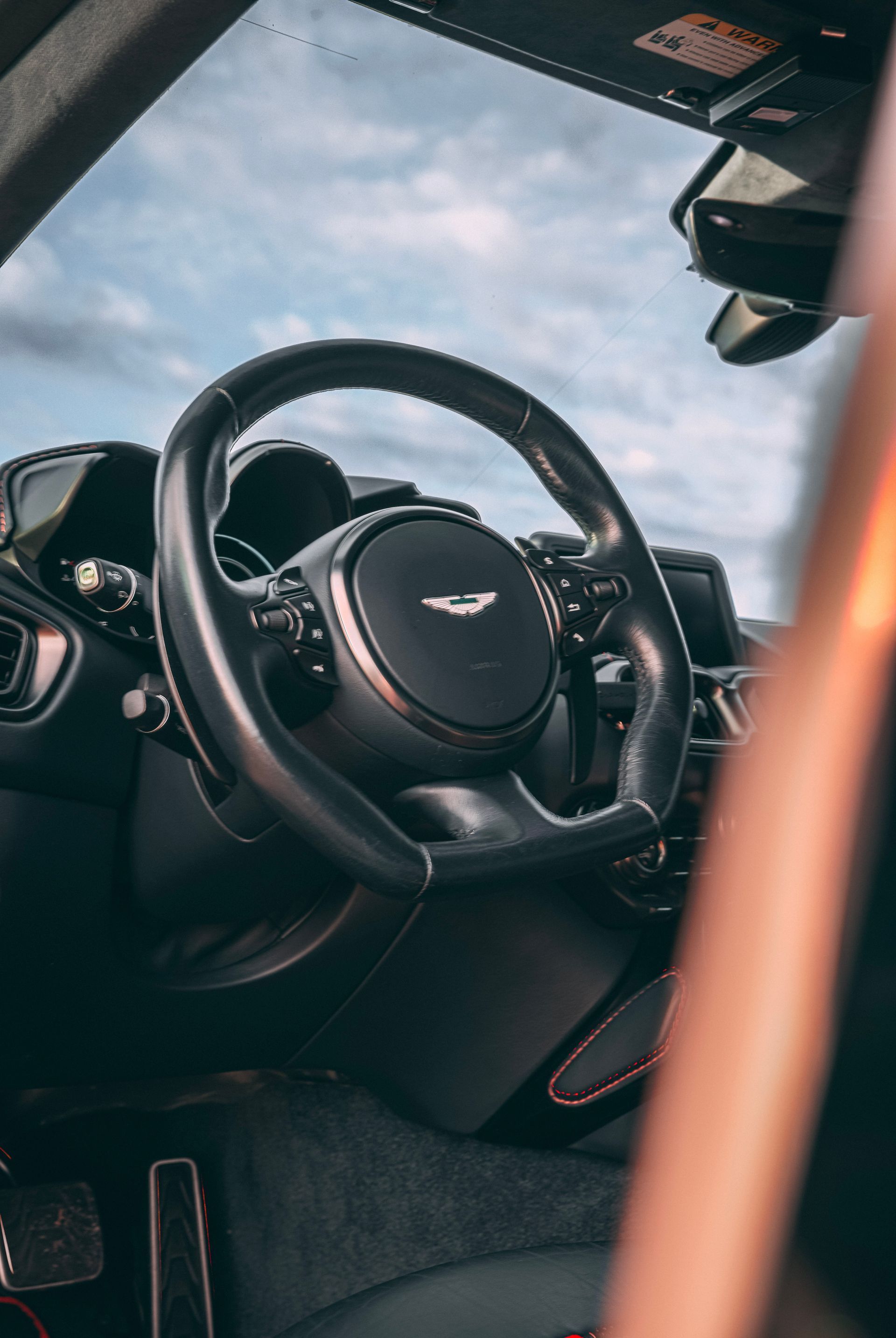 Black Aston Martin steering wheel with controls and logo, dark interior, against sky.