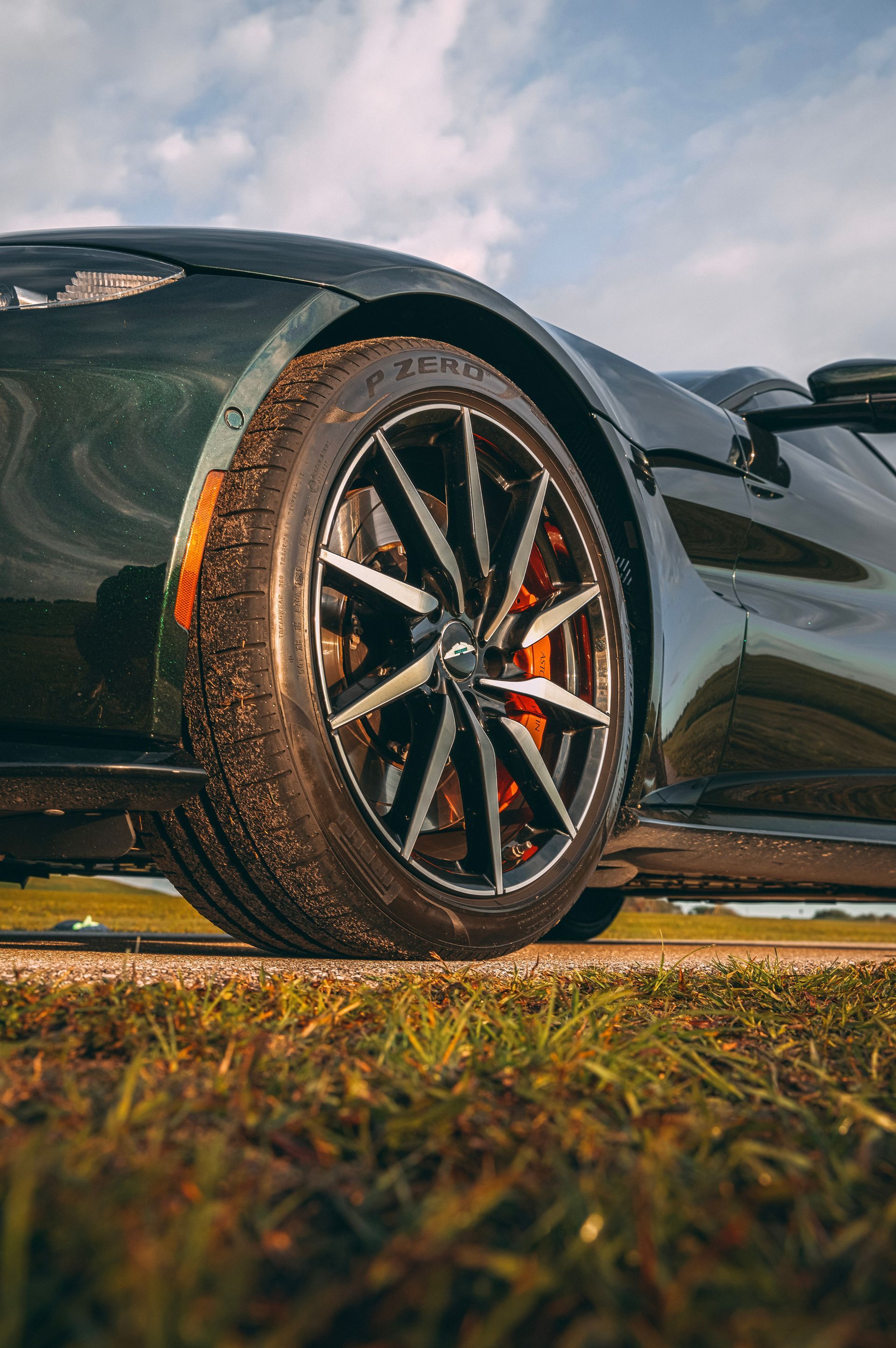 Dark green sports car's front wheel on a grassy verge, showing the tire, black rim, and red brake caliper, against a blue sky.