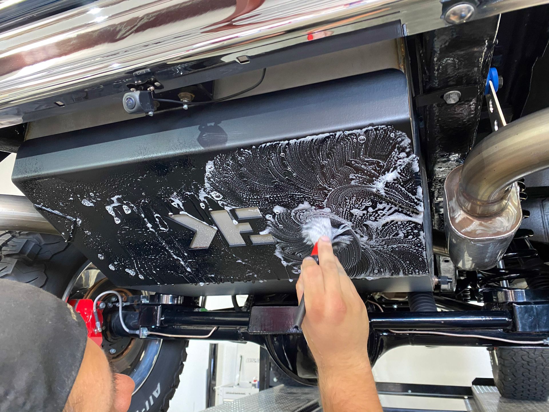 Person cleaning a black metal car part with a brush, revealing a silver logo.