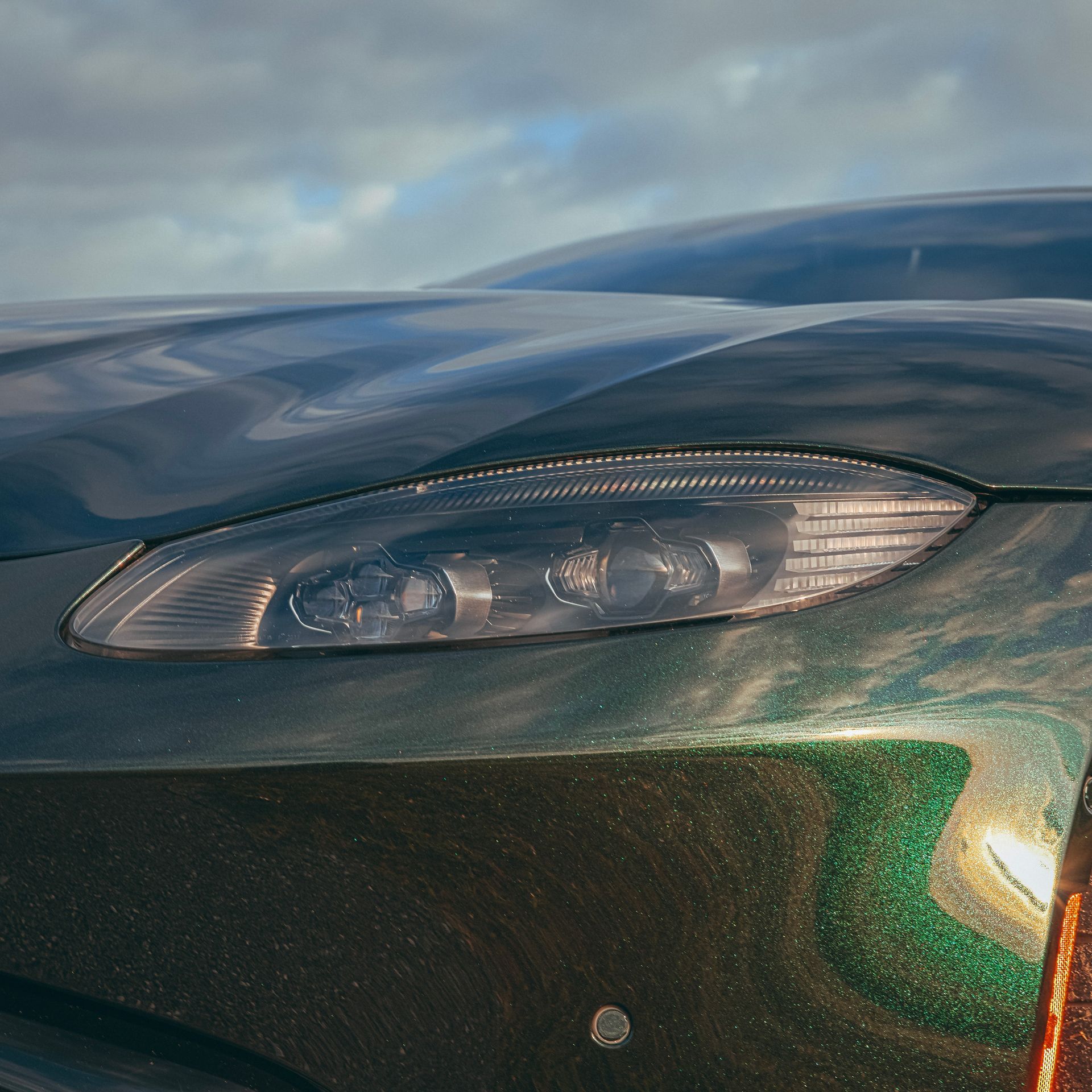 Close-up of a dark green luxury car's front with a headlight, reflecting sunlight.