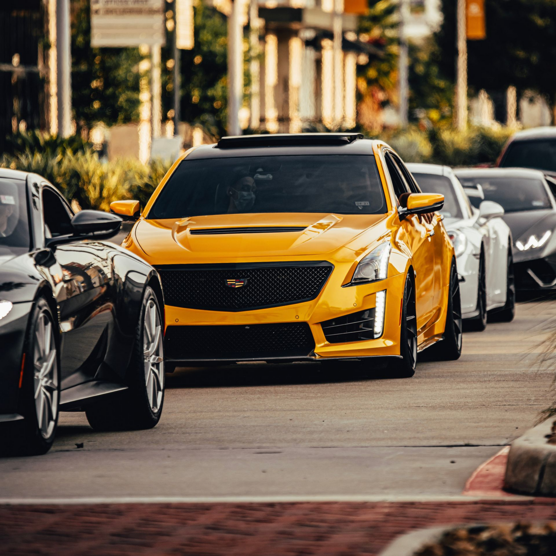 Line of sports cars, including a yellow Cadillac, driving down a street lined with shops.