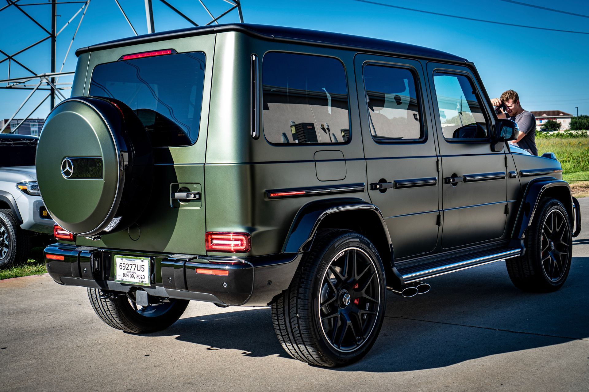 Green Mercedes-Benz G-Class SUV, spare tire on back, parked on asphalt, person taking a photo.
