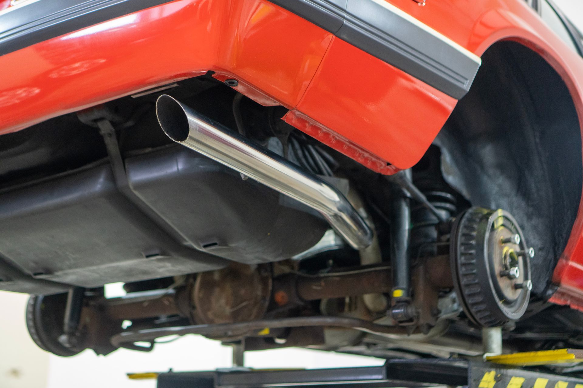 Red car's underside with exhaust pipe, gas tank, suspension, and rear wheel on a lift.