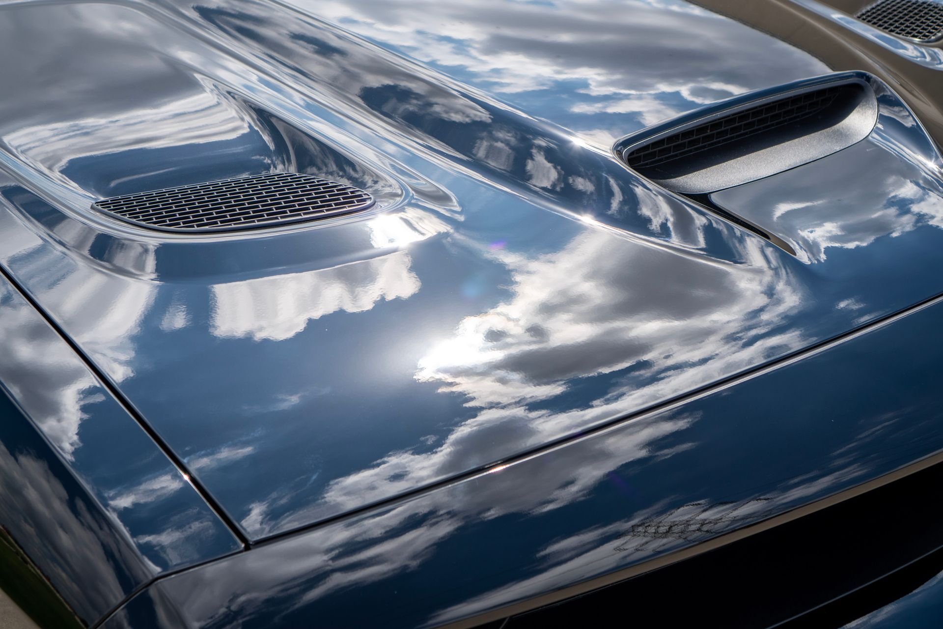 Reflective, dark blue car hood with sky and clouds visible in the polished surface.