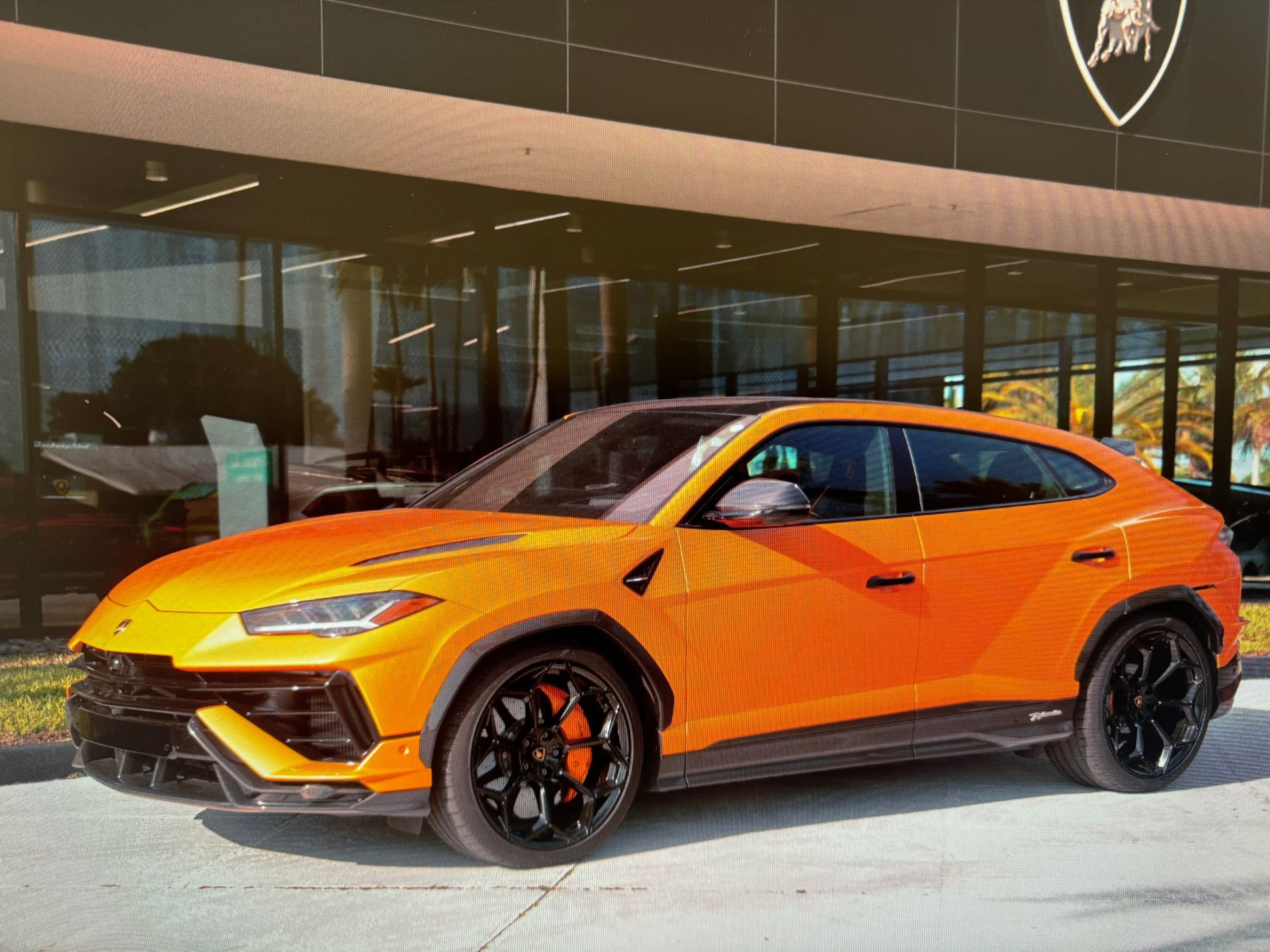 Bright orange Lamborghini Urus SUV parked in front of a dealership. Black rims and brake calipers.