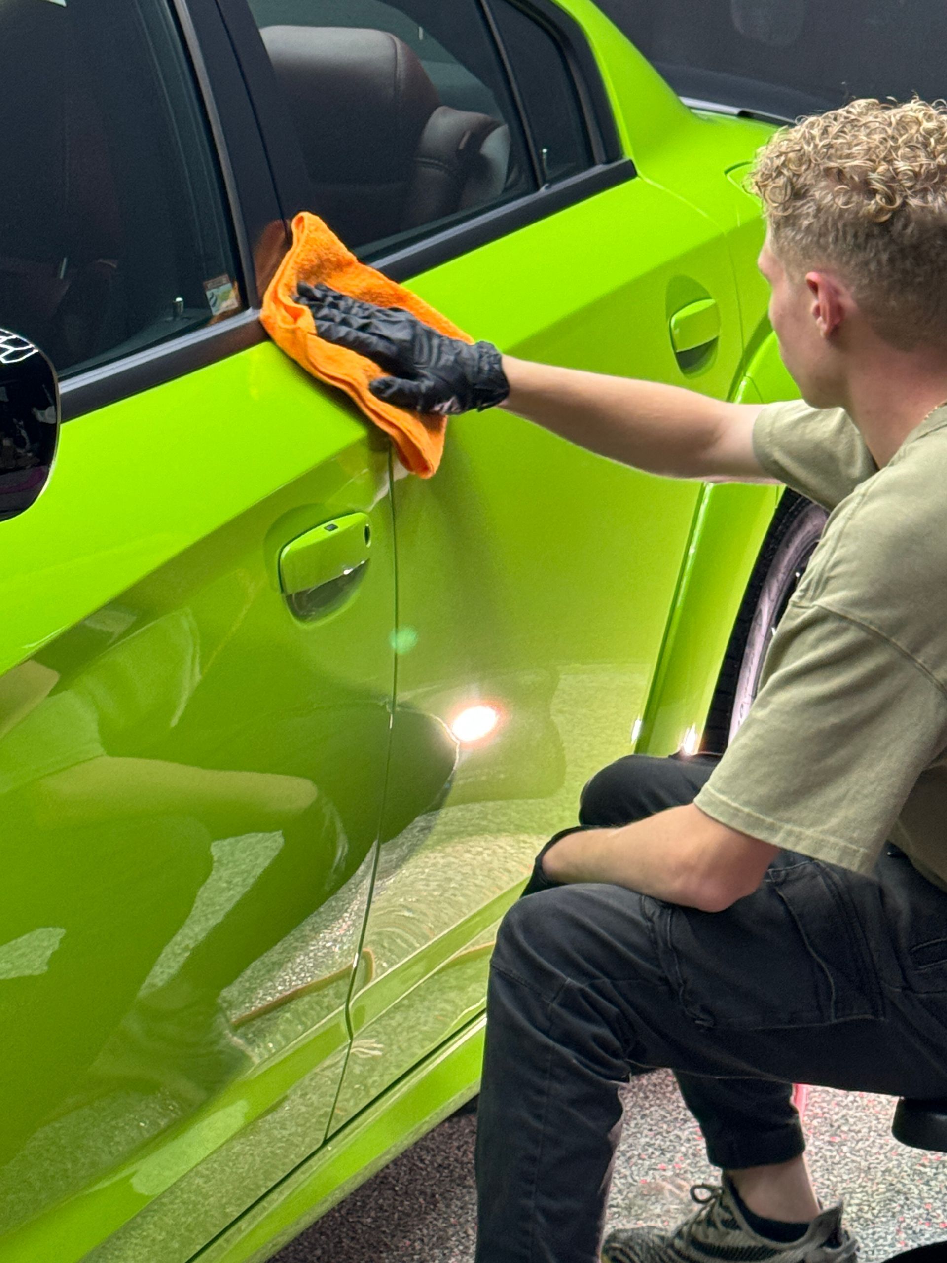 Person in black gloves wiping down a lime green car with an orange cloth.